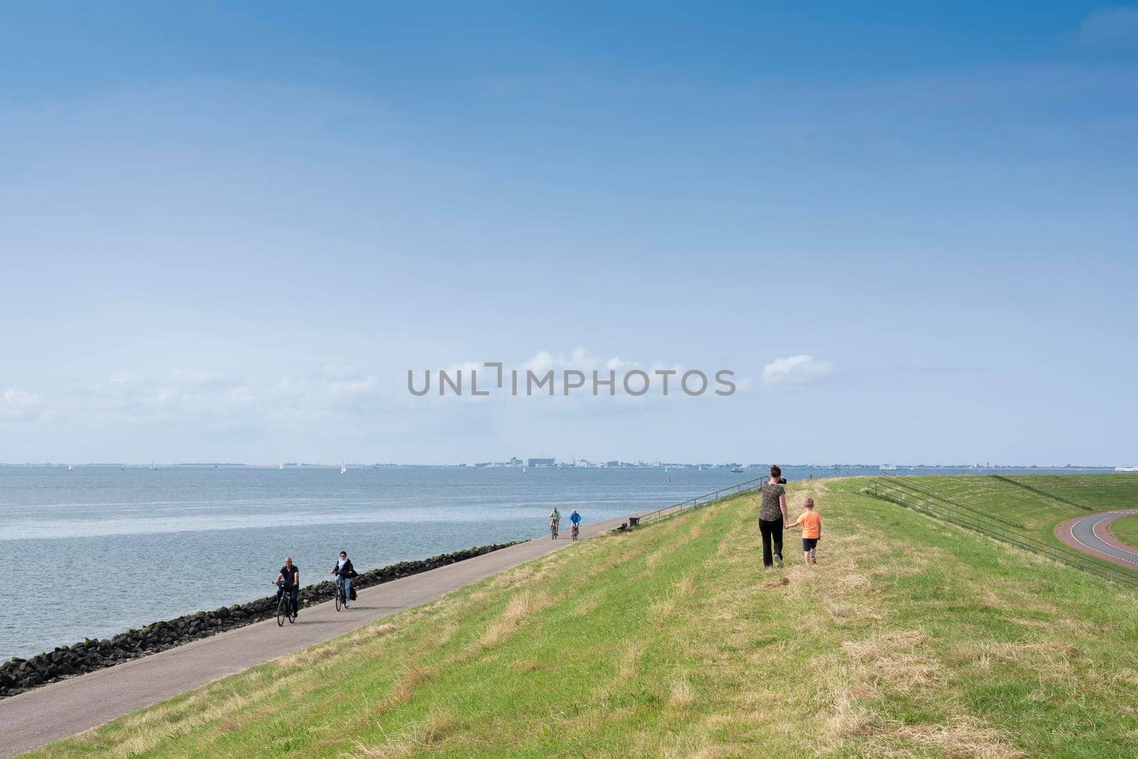 people walk and ride bicycle on dike near oudeschild on the dutch island of texel under blue sky in summer by ahavelaar