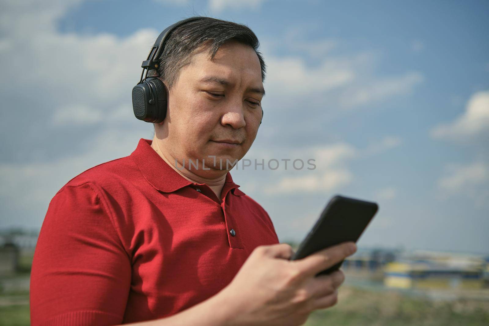 Middle-aged Asian man in headphones outdoors listening to music against the background of the sky, mobile phone in his hand