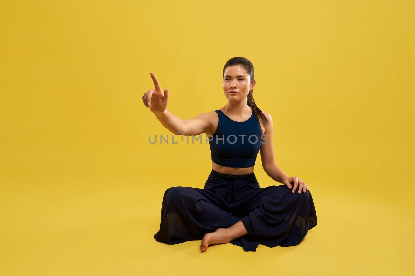 Front view of sporty girl sitting with crossed legs on floor, raising hand, holding finger up, looking forward. Slim brunette doing yoga. Isolated on yellow studio background.
