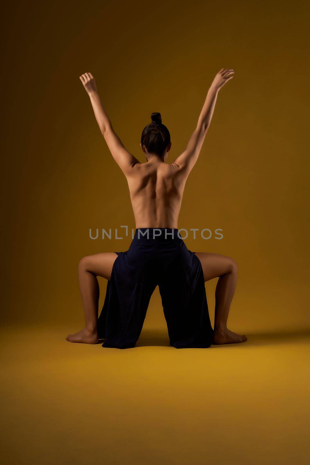 Back view of slim girl with hair bun standing with legs bent and hands raised. Young female with bare back stretching, yoga practicing. Isolated on yellow studio background.