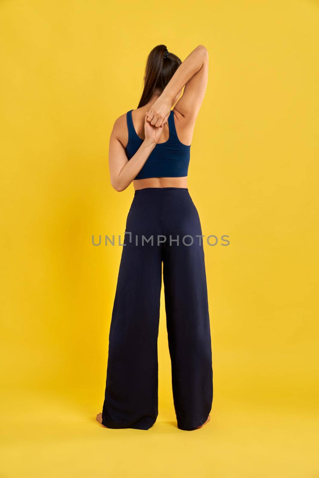 Back view of flexible girl with pony tail standing backwards, holding hands in lock. Spoert fmale stretching, yoga doing, meditating. Isolated on yellow studio background.