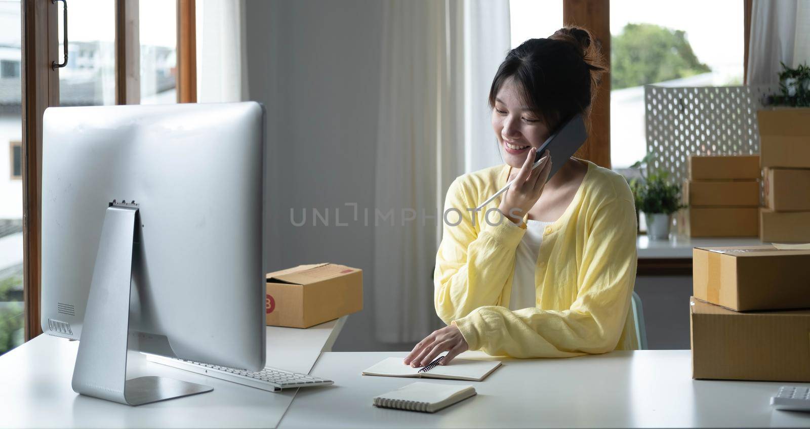 A portrait of Asian woman, e-commerce employee sitting in the office full of packages on the table using a laptop and smartphone, for SME business, e-commerce, technology and delivery business. by wichayada