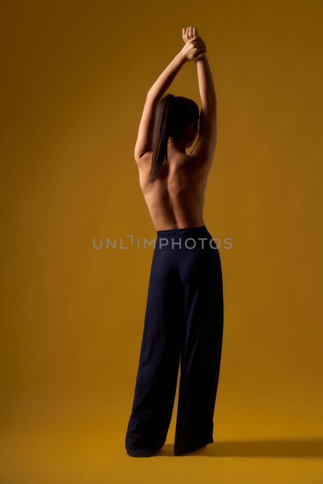 Back view of female with pony tail doing yoga indoors. Flexible woman with bare back standing backwards, raising hands, meditating. Concpt oof yoga doing