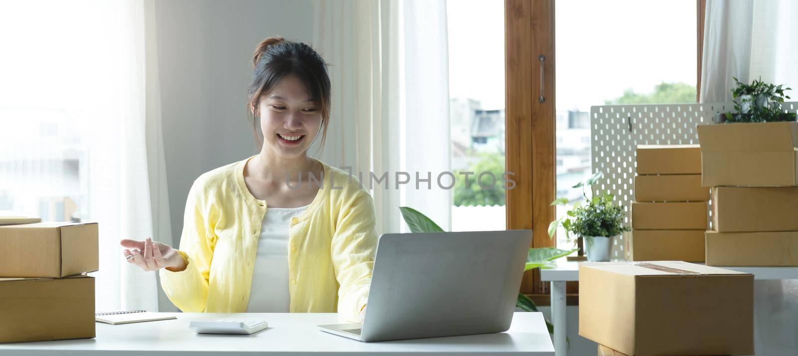 A portrait of Asian woman, e-commerce employee sitting in the office full of packages on the table using a laptop and calculator, for SME business, e-commerce, technology and delivery business..