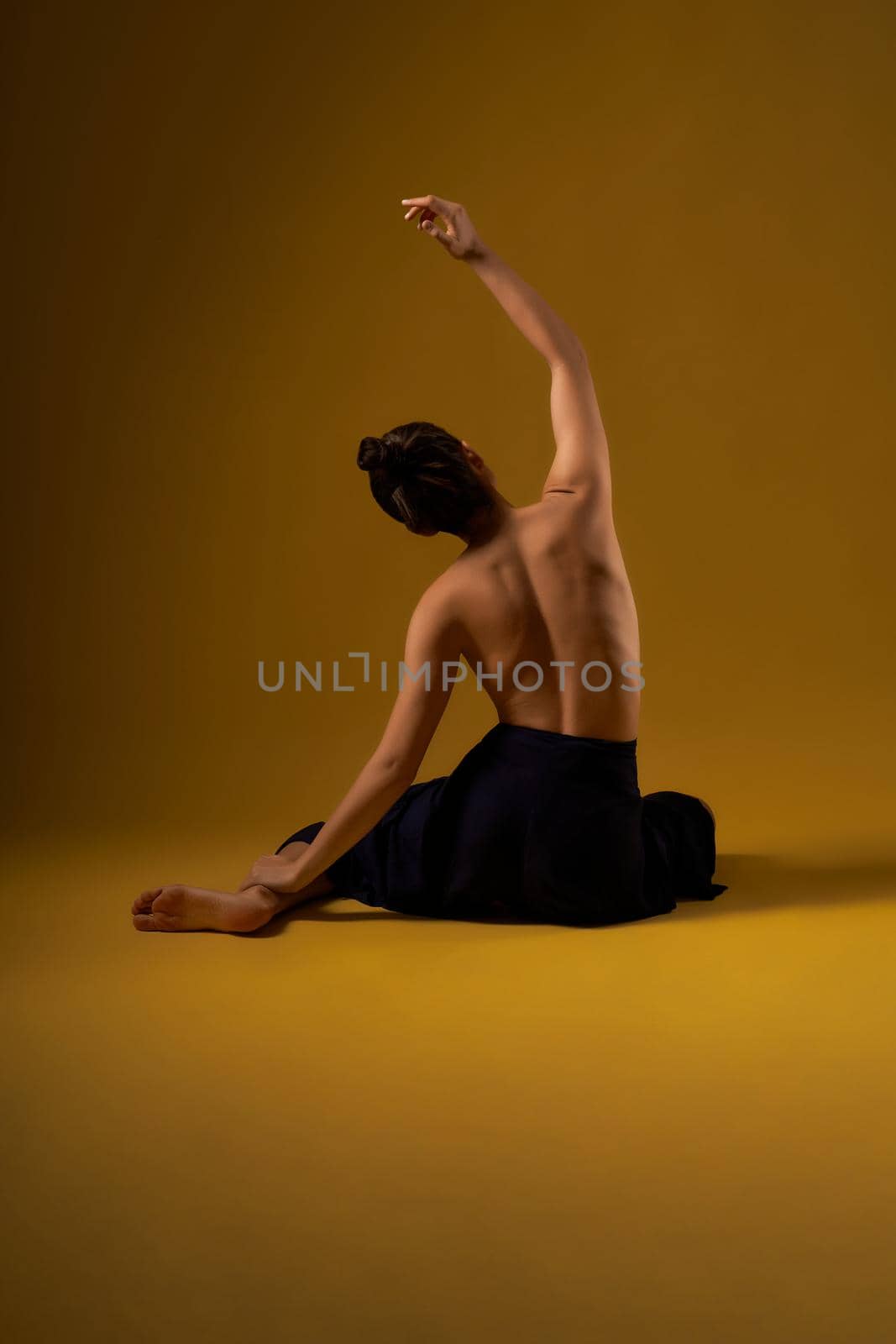Back view of flexible girl with bare back sitting backwards on floor, with raised hand. Brunette with hair bun stretching, meditating. Concept of new age and yoga practicing.