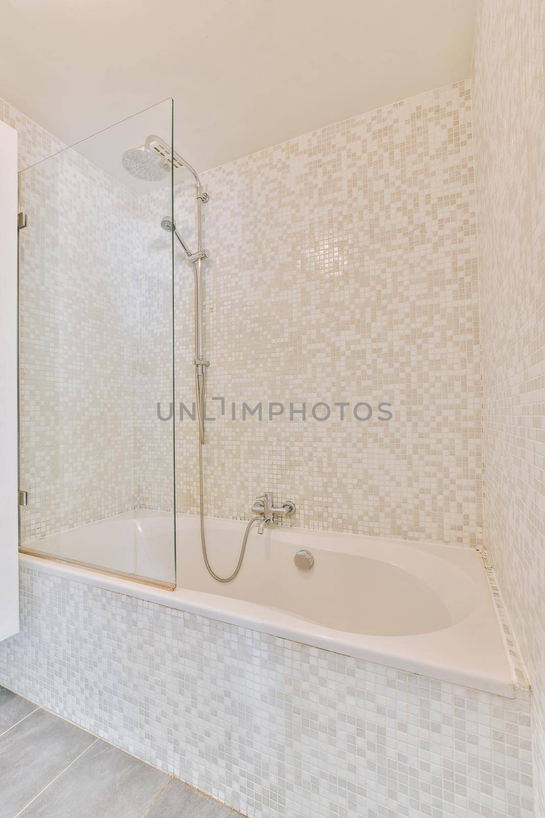 Bathtub with shower and glass partition placed in corner in small light bathroom with beige tile walls