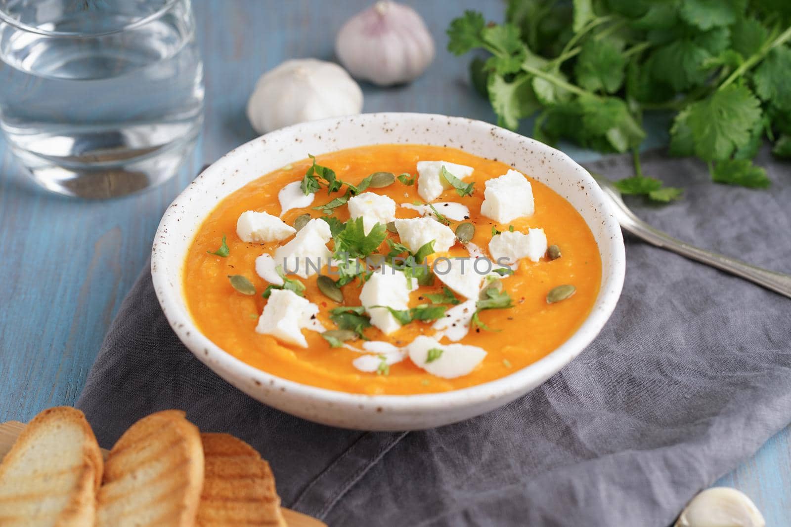 Pupmkin cream soup puree with ricotta, dietary vegetarian lunch on blue wooden table, side view. Glass of water, toast, parsley, garlic.
