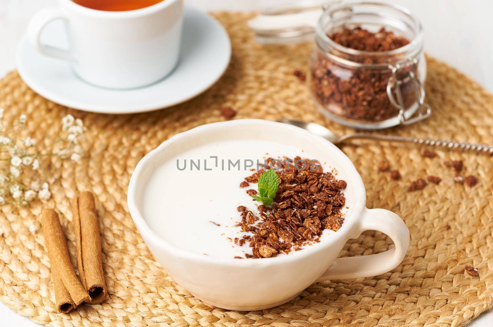 Yogurt with chocolate granola in cup, breakfast with tea on beige background, side view