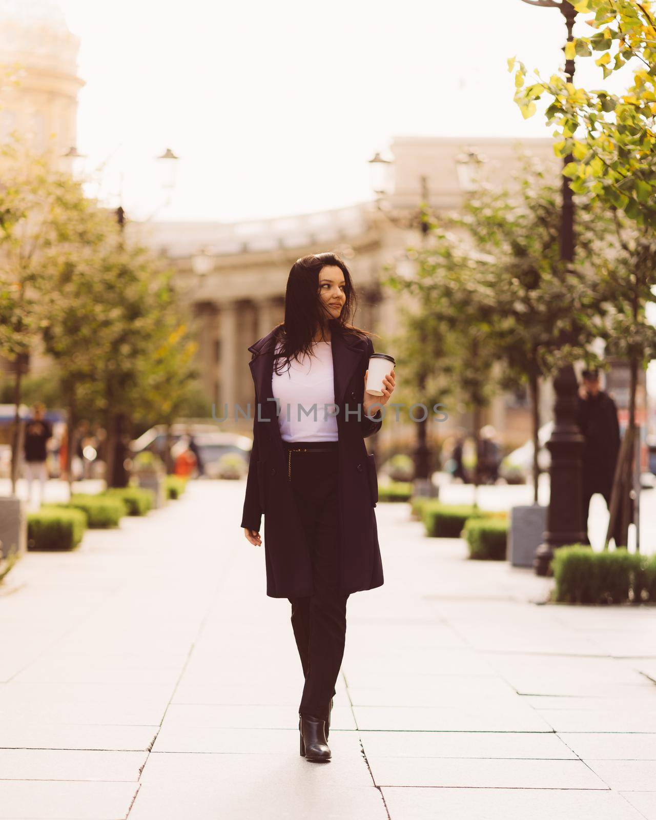 Beautiful serious smart brunette girl holding cup of coffee in hands goes walking down street of St. Petersburg in city center. Charming thoughtful woman with a long dark hair wanders alone