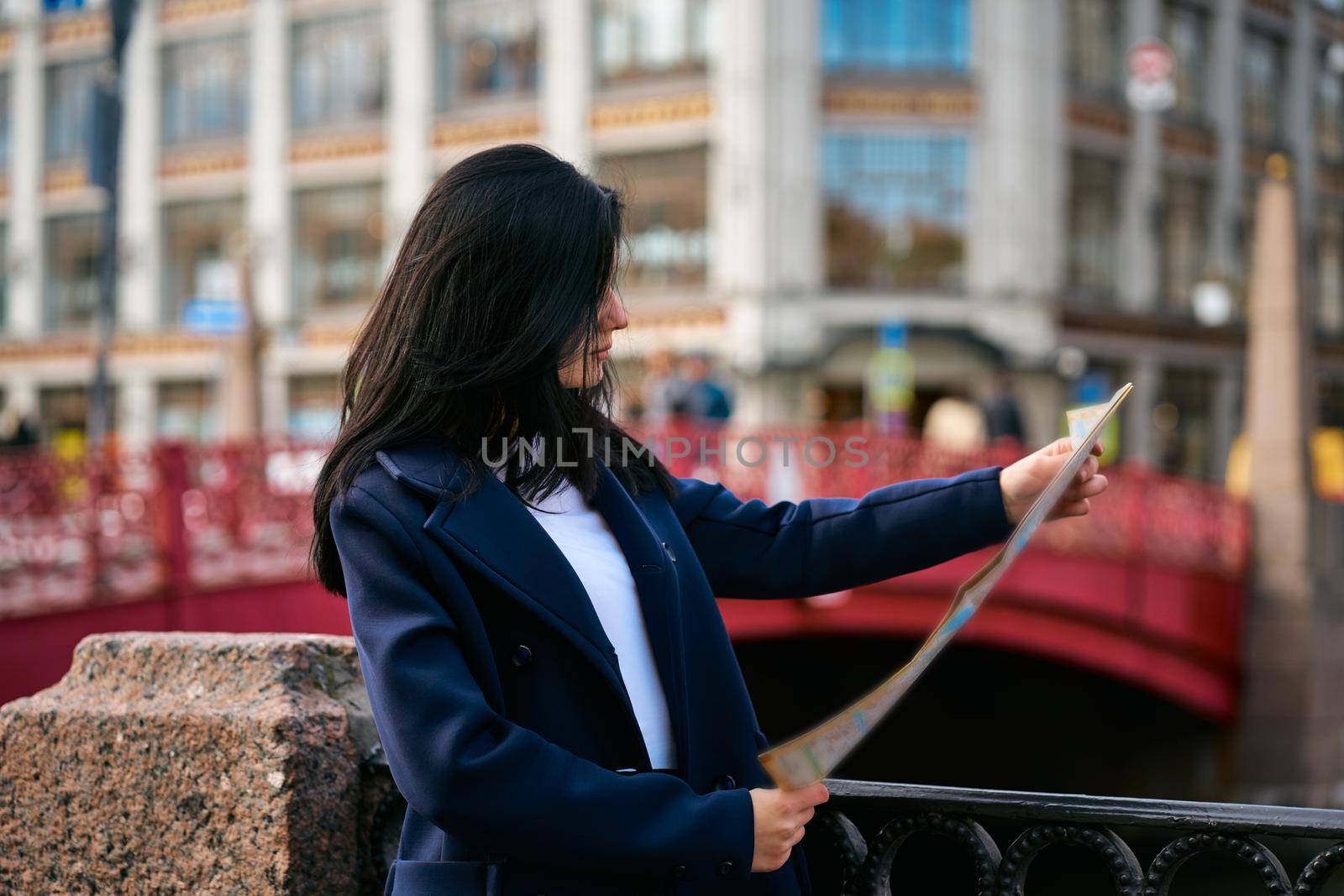 Young woman standing on waterfront of big city and looking at guide, a tourist looking for for attractions. A charming thoughtful fashionably dressed woman with long dark hair travels through Europe