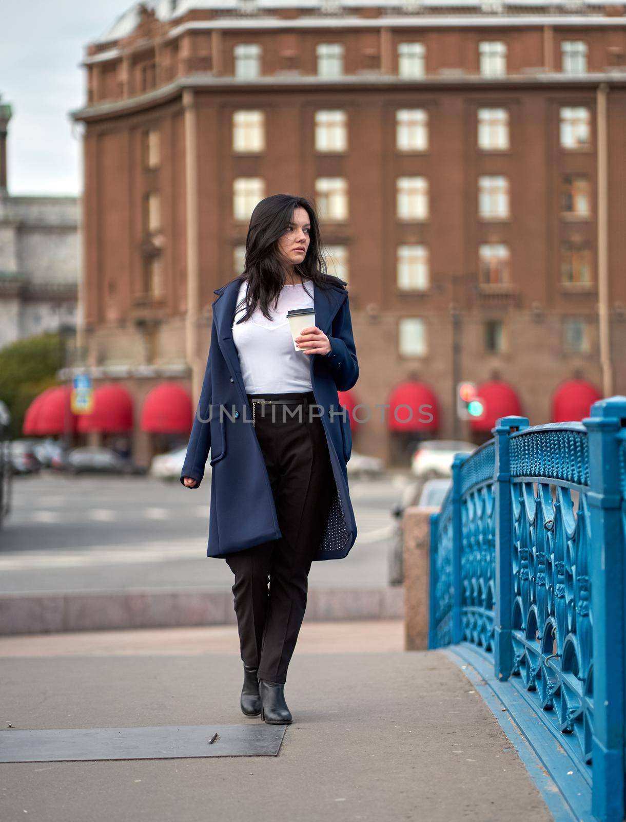 Beautiful serious smart brunette girl holding cup of coffee in hands goes walking down street of St. Petersburg in city center on a bridge. Charming thoughtful woman with long dark hair wanders alone