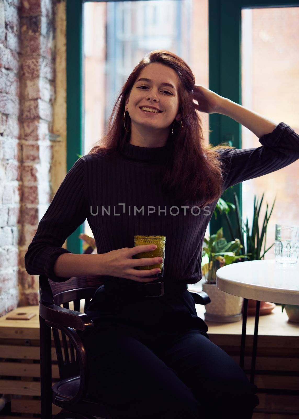 Beautiful serious stylish fashionable smart girl sits by window in cafe, smiles beautifully and drinks healthy vegan smoothie or latte. Charming thoughtful woman with long dark brown hair by NataBene