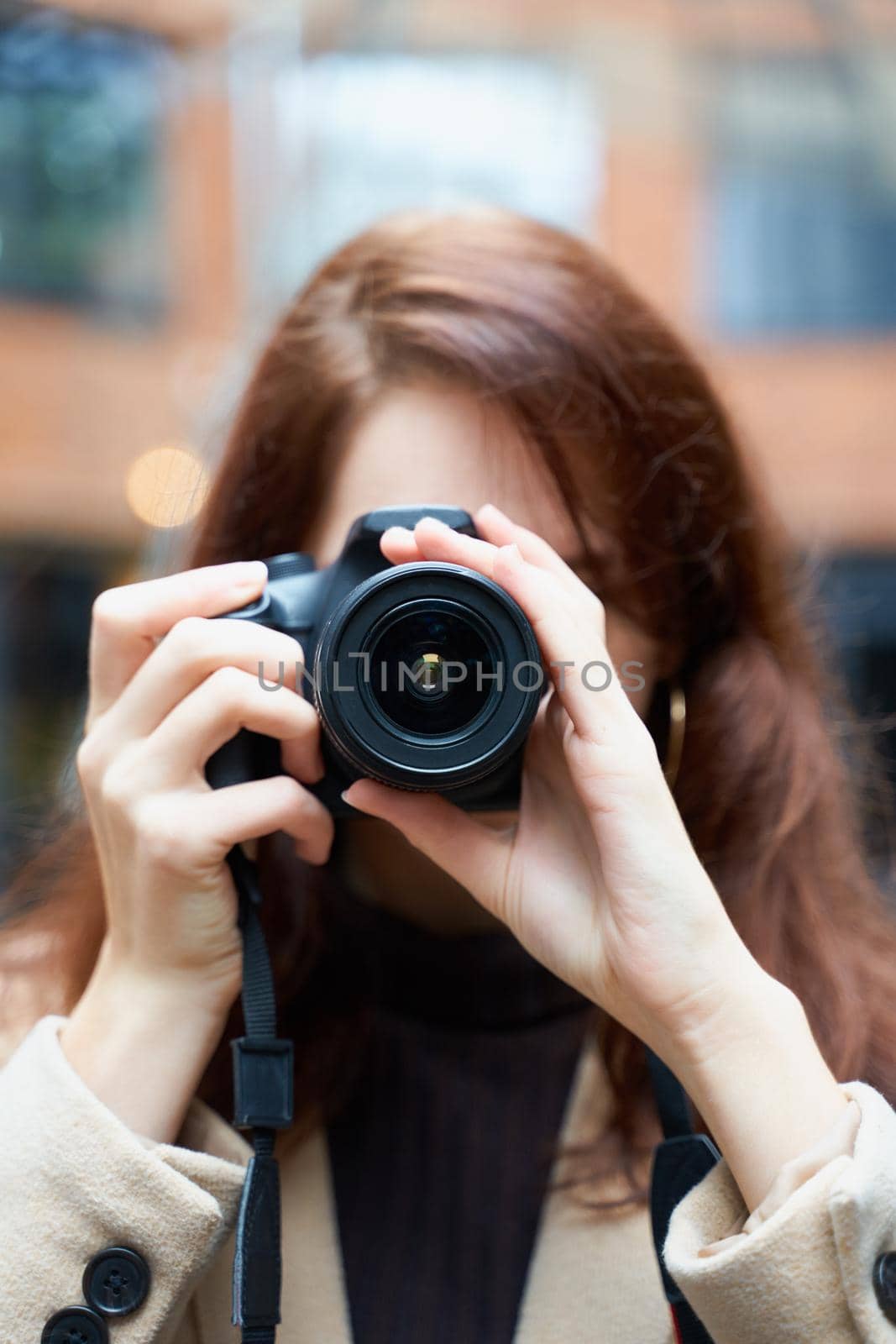 Selective focuson lens. Beautiful stylish fashionable girl holds camera in her hands and takes pictures. Woman photographer with long dark hair in a city, urban shoot, vertical. unrecognizable person