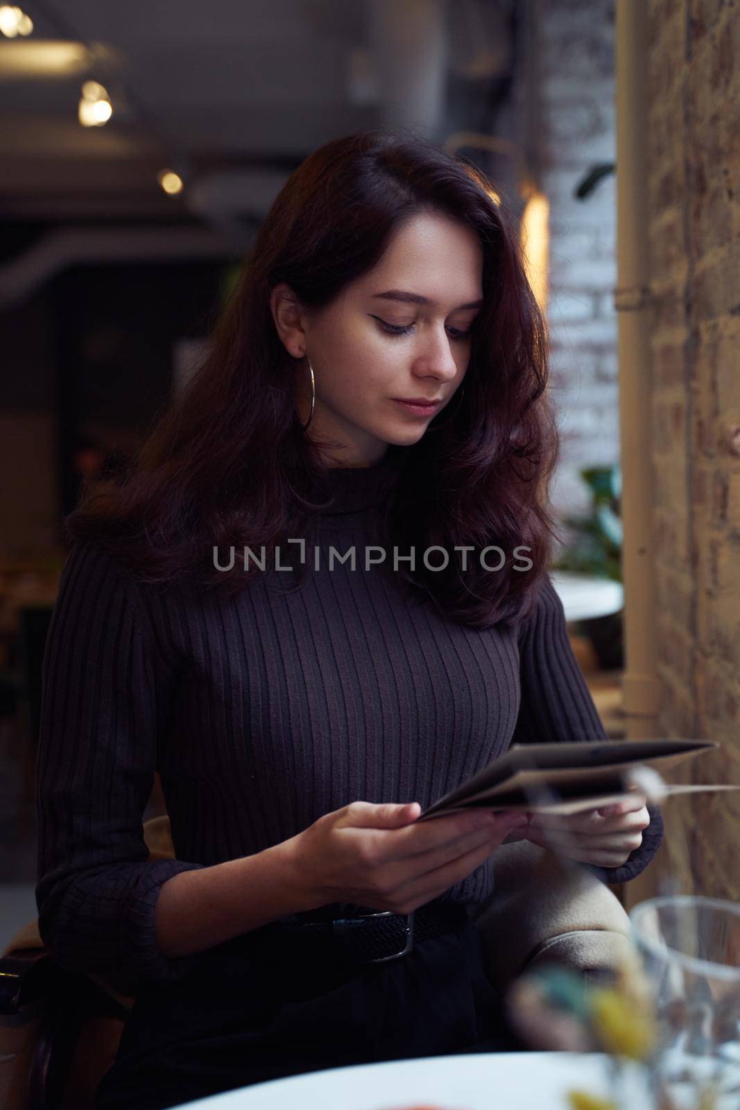 Beautiful serious stylish fashionable smart girl sit in cafe in loft style on lunch. Ready to place an order, studying menu. Charming thoughtful woman with long dark brown hair, vertical, moody