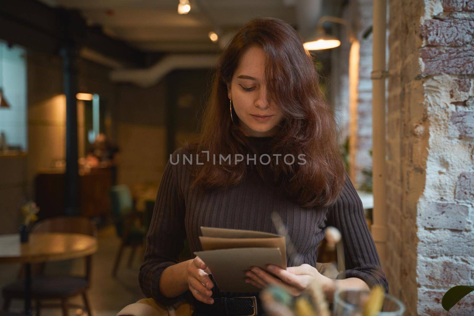 Beautiful serious stylish fashionable smart girl sit in cafe in loft style on lunch. Ready to place an order, studying menu. Charming thoughtful woman with long dark brown hair.