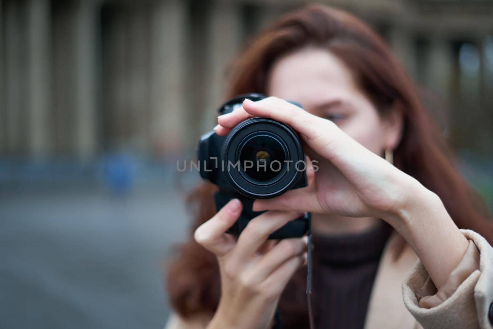 Selective focus on lens. Beautiful stylish fashionable girl holds camera in hands and takes pictures. Woman photographer with long dark hair in city, urban shoot, vertical. unrecognizable person, copy space