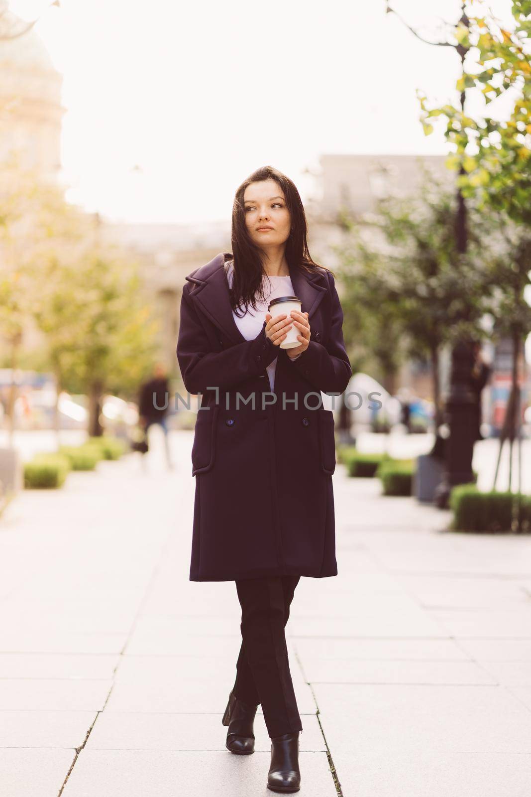 Beautiful serious smart brunette girl holding cup of coffee in hands goes walking down street of St. Petersburg in city center. Charming thoughtful woman with a long dark hair wanders alone, vertical