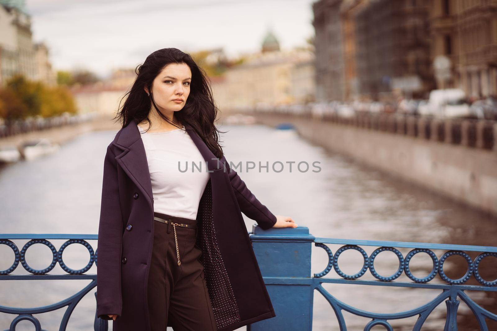 Charming thoughtful fashionably dressed woman with long dark hair travels through Europe, standing in city center of St. Petersburg. A beautiful girl wanders alone through autumn streets, copy space