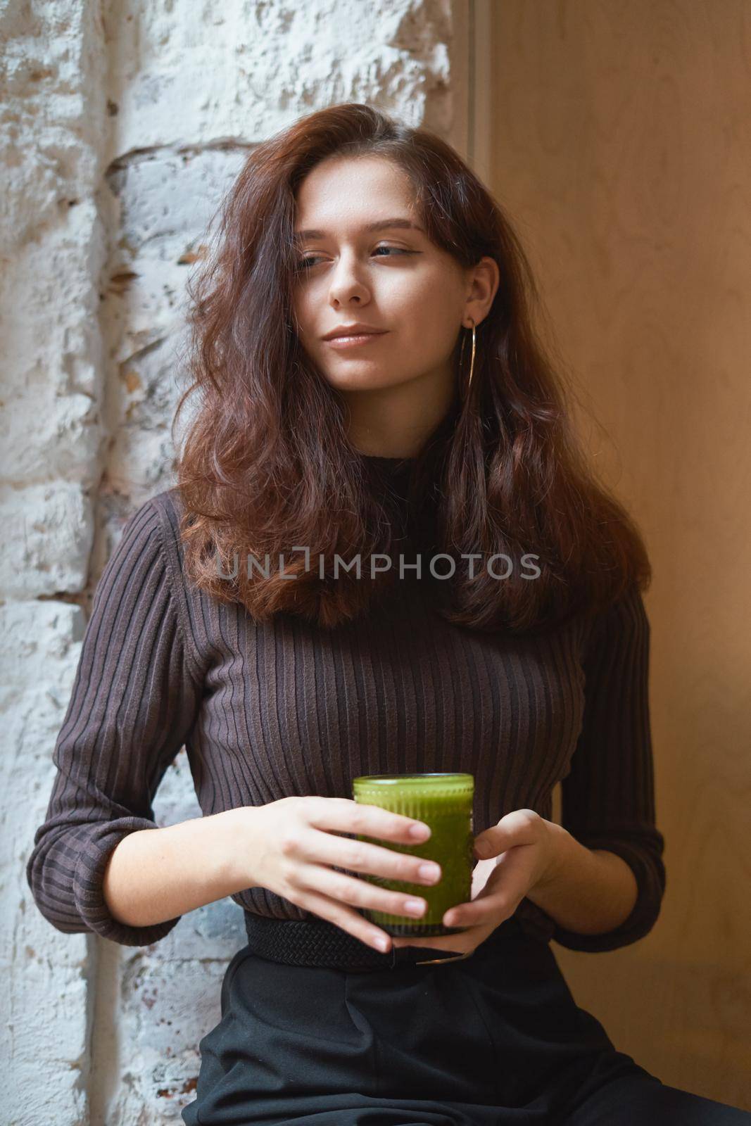 Beautiful serious stylish fashionable smart girl is sitting in cafe and drinking healthy green smoothie or latte vegan. Charming thoughtful woman with long dark brown hair, vertical