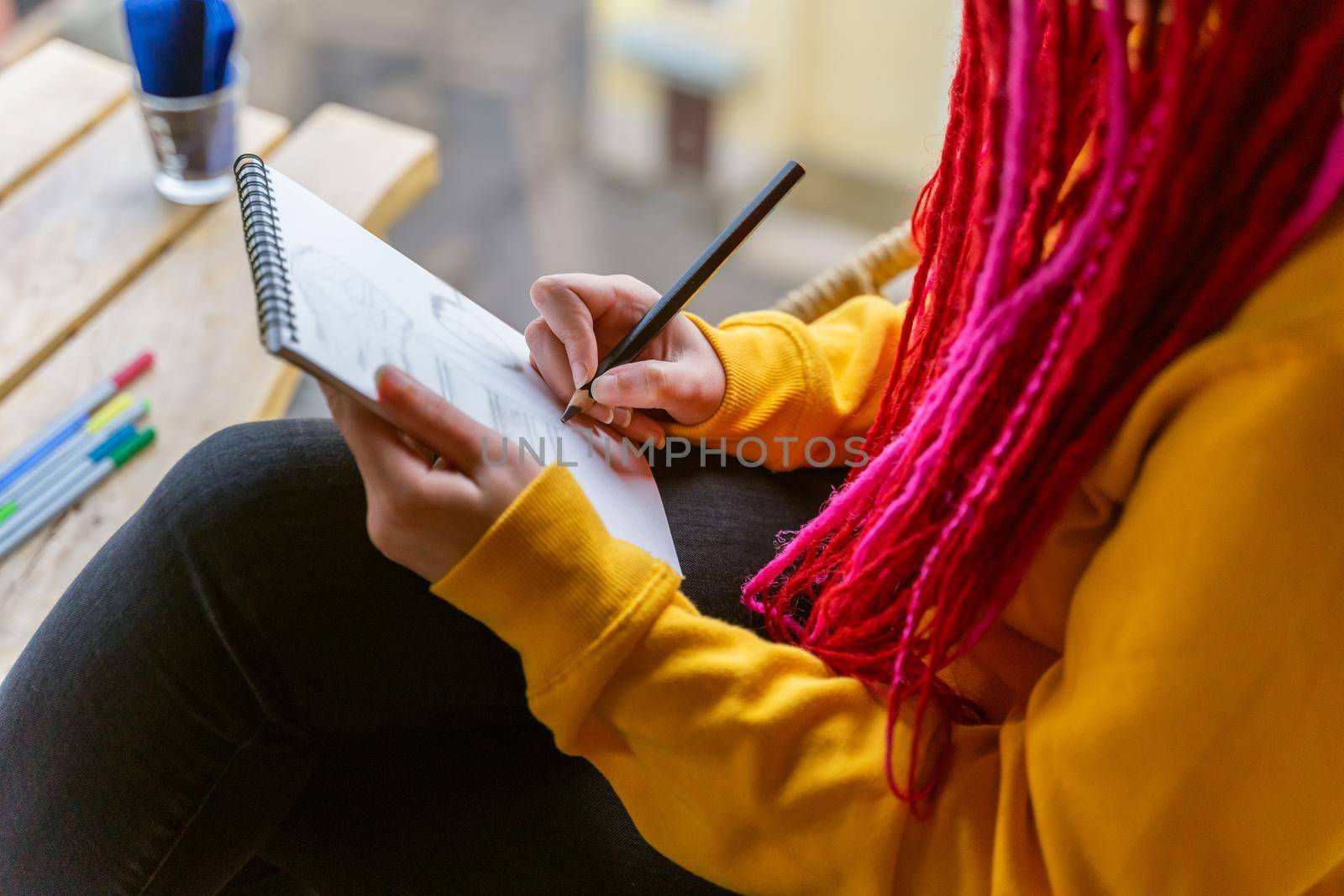 Unrecognizable person, girl artist, illustrator draws in notebook, makes sketch. Close-up of woman with long pink dreadlocks in informal setting, in casual comfortable clothes sitting in a cafe
