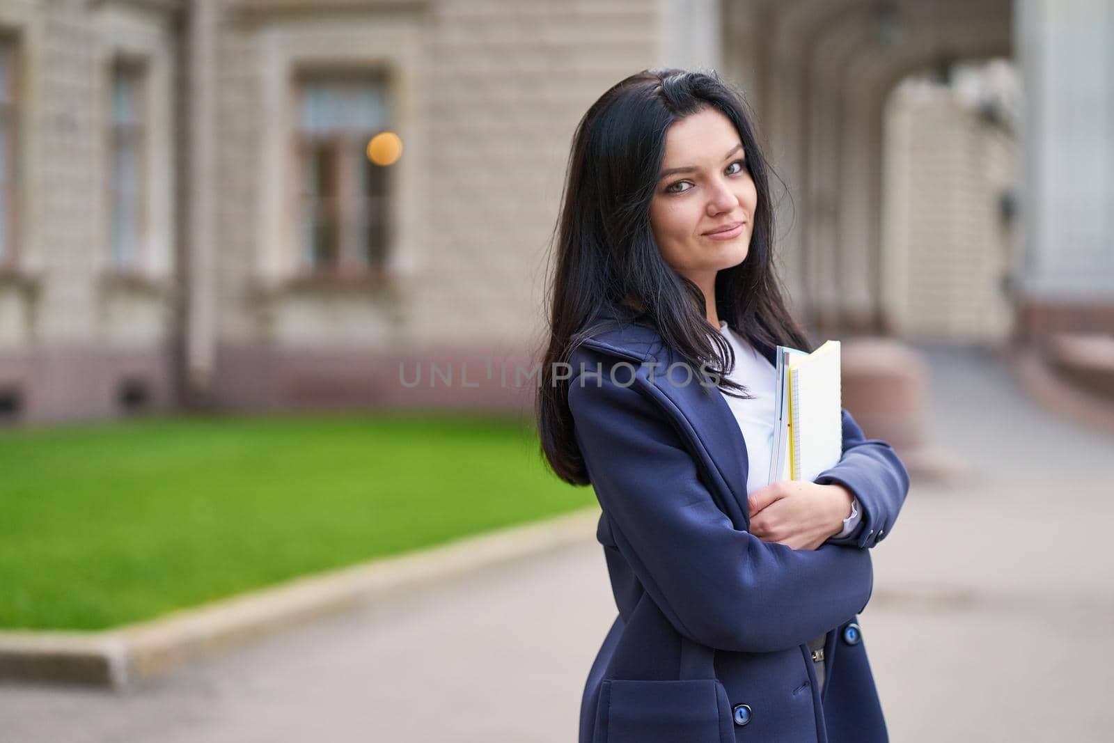 Smiling girl brunette student holding notebooks and textbooks, stands at the University on the street of St. Petersburg. A charming woman with long dark hair is studying at course, getting education by NataBene