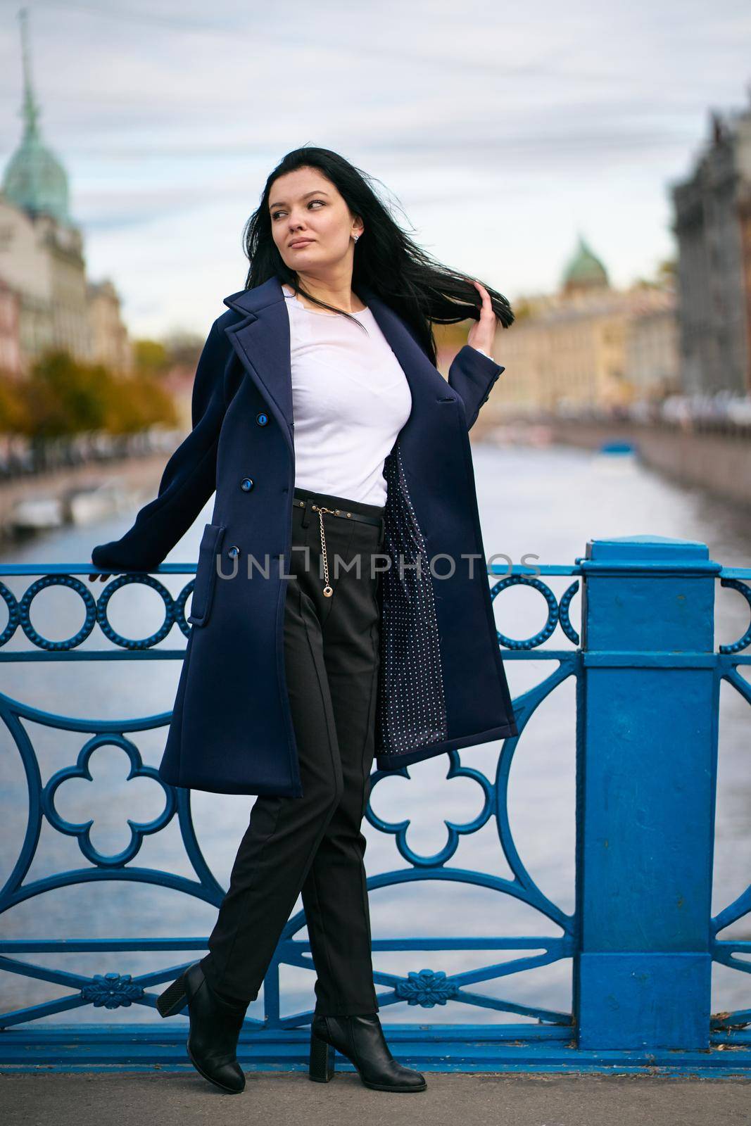 Charming thoughtful fashionably dressed woman with long dark hair travels through Europe, standing in city center of St. Petersburg. A beautiful girl wanders alone through autumn streets by NataBene