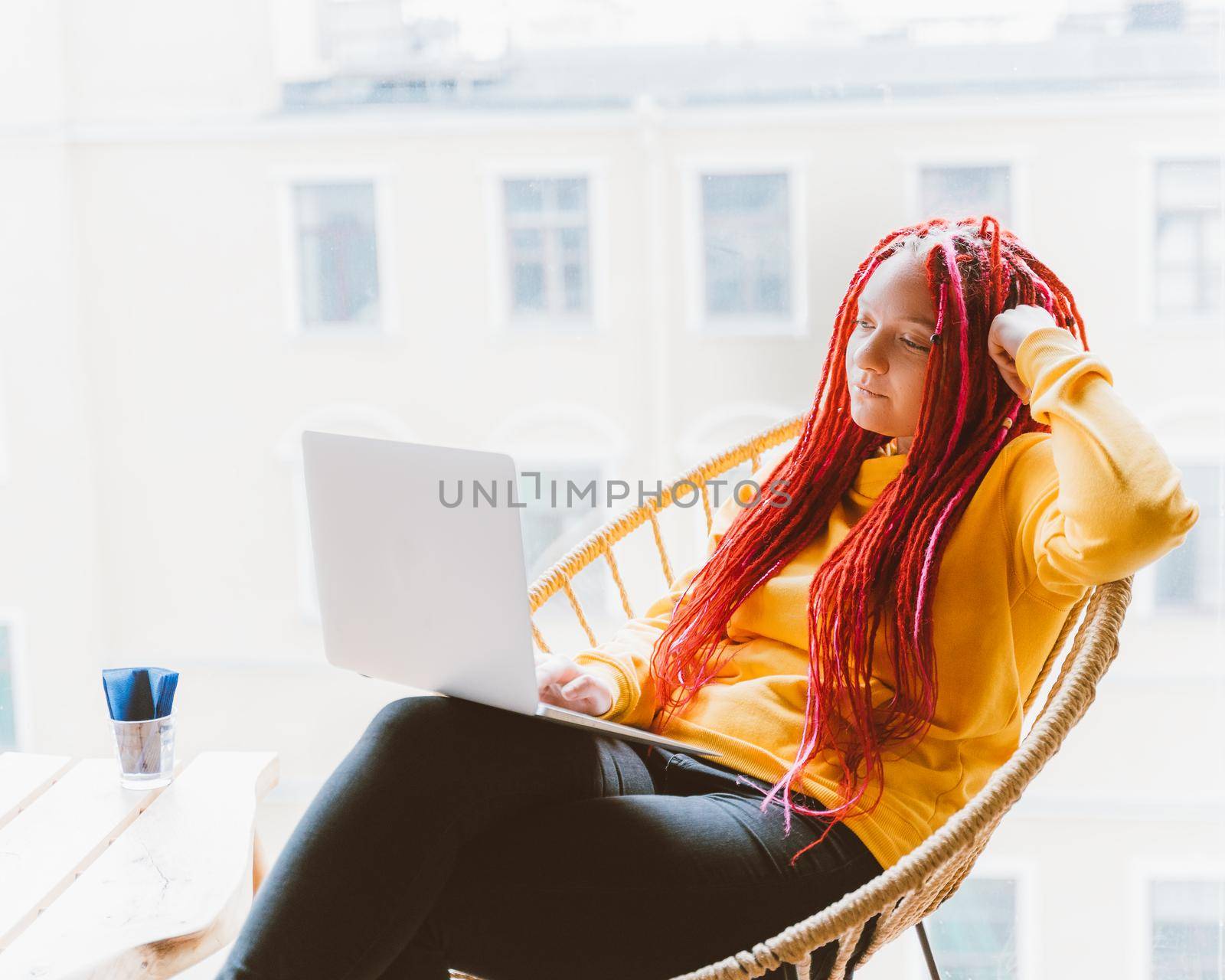 Digital nomad concept. Girl freelancer remotely working on a laptop in a cafe, coworking. Woman with long pink dreadlocks in an informal setting, in casual comfortable clothes sitting at chair