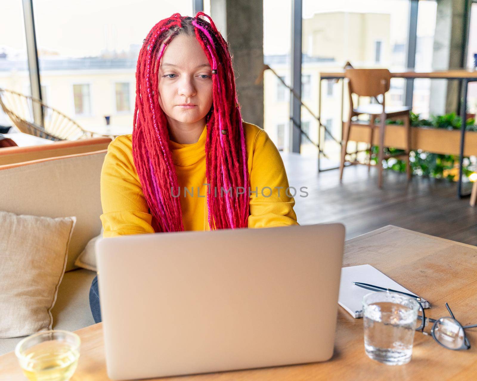 Digital nomad concept. Girl freelancer remotely working on a laptop in a cafe, coworking. Woman with long pink dreadlocks in an informal setting, in casual comfortable clothes sitting at the table.