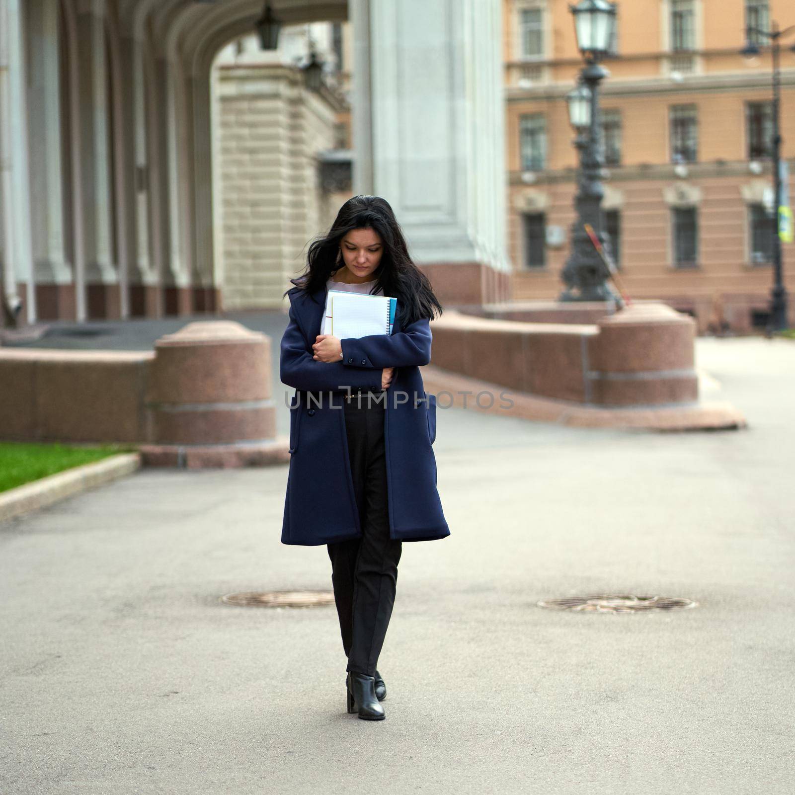 Beautiful serious smart girl brunette student holding notebooks and textbooks, goes walking at University on street of St. Petersburg. A charming woman with long dark hair is studying at course by NataBene