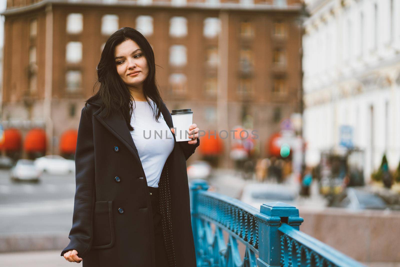 Beautiful serious smart brunette girl holding cup of coffee in hands goes walking down street in a enter on blue bridge. Charming thoughtful woman with long hair wanders alone, immersed in thoughts