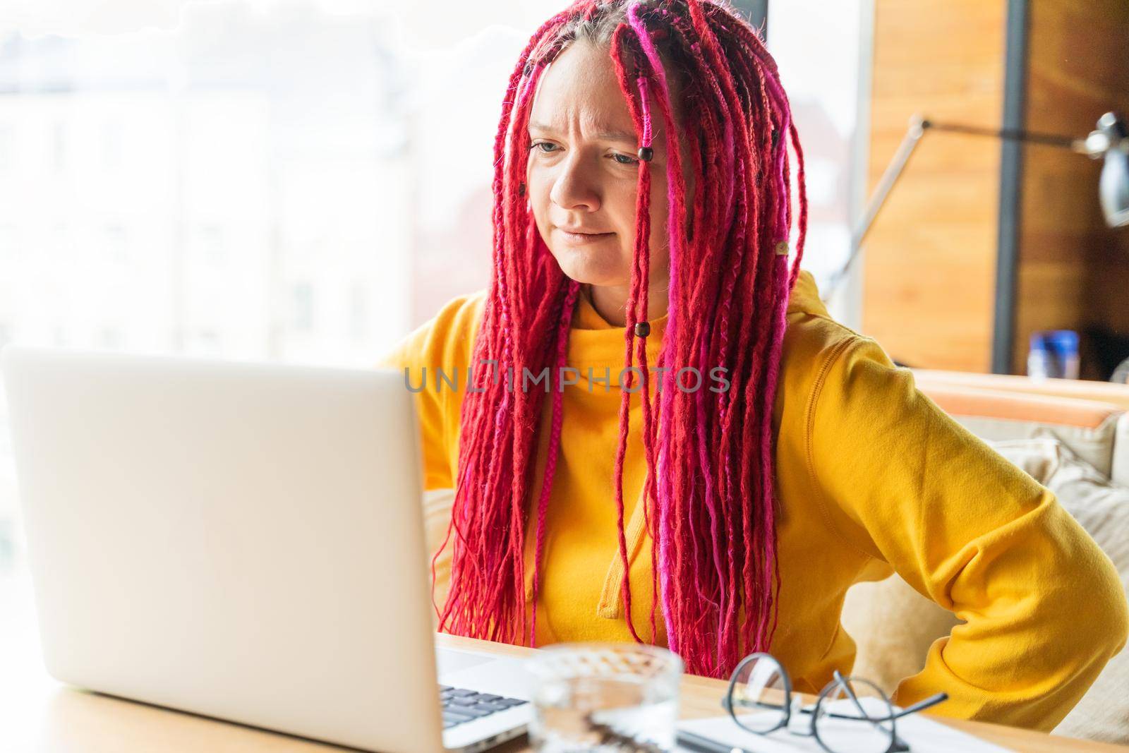 Woman looks at a laptop and gets angry, surprised, annoyed. Girl with a long pink hair, designer, creative specialist, freelancer. Difficulties in understanding, in work, in online communication