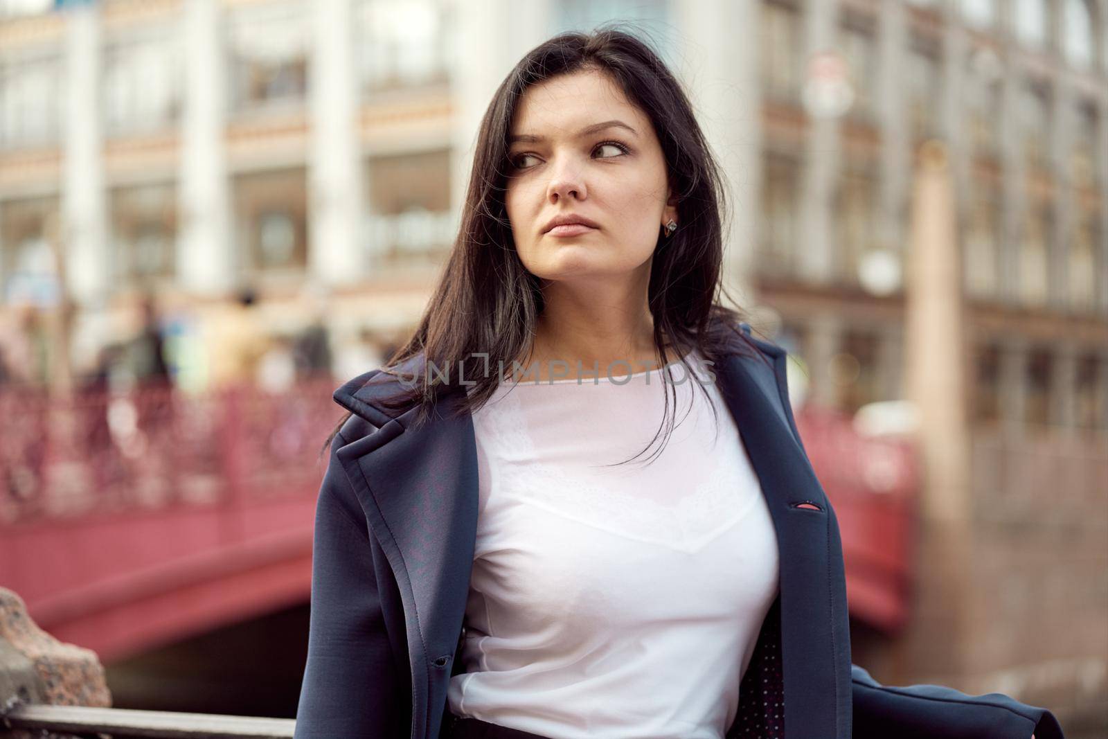 Beautiful serious smart brunette girl stands on the bridge on the street of St. Petersburg in the city center. A charming thoughtful fashionably dressed woman with long dark hair looks away, copy space