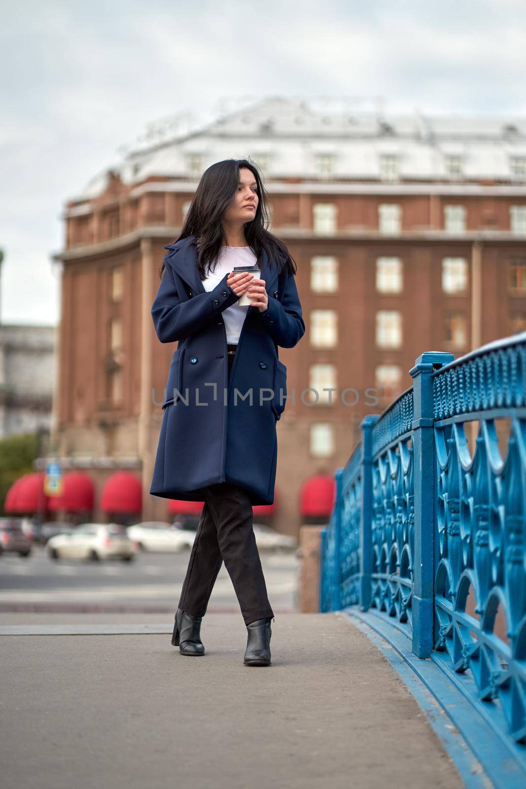 Beautiful serious smart brunette girl holding cup of coffee in hands goes walking down street of St. Petersburg in city center on bridge. Charming thoughtful woman with long dark hair, vertical