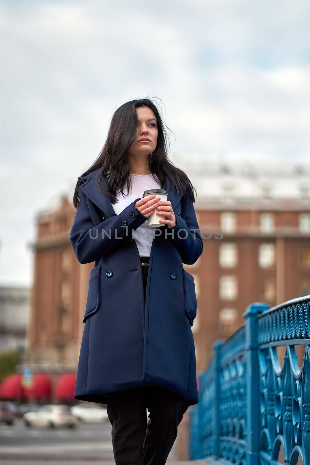 Beautiful serious smart brunette girl holding cup of coffee in hands goes walking down street of St. Petersburg in city center on bridge. Charming thoughtful woman with long dark hair wanders alone by NataBene