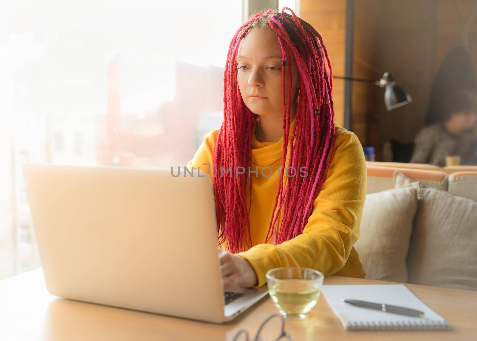 Digital nomad concept. Girl freelancer remotely working on a laptop in a cafe, coworking. Woman with long pink dreadlocks in an informal setting, in casual comfortable clothes sitting at the table.