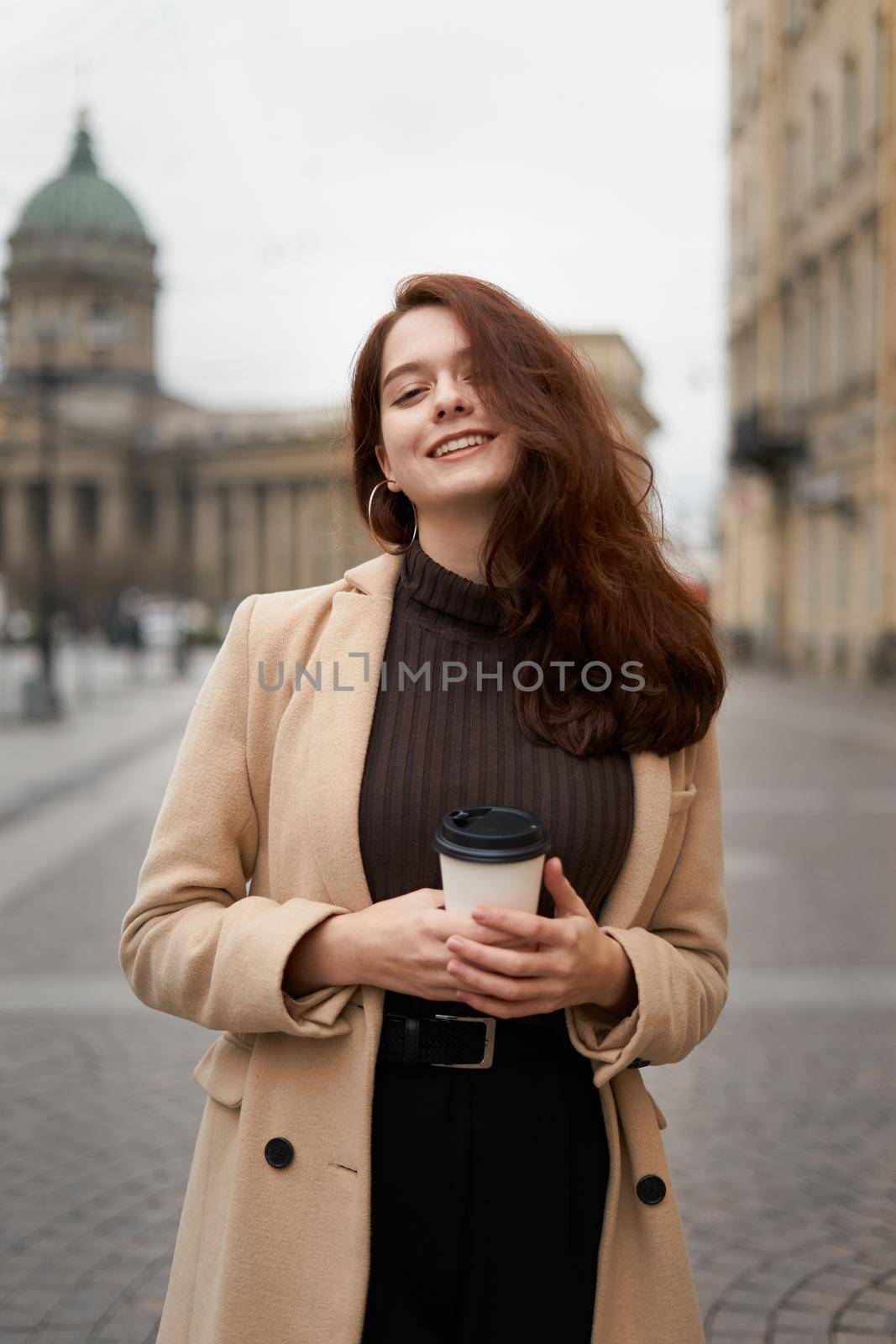 Beautiful serious stylish fashionable smart girl holding cup of coffee in hands and smiles, goes walking down street of St. Petersburg in city center. Charming thoughtful woman with long dark hair