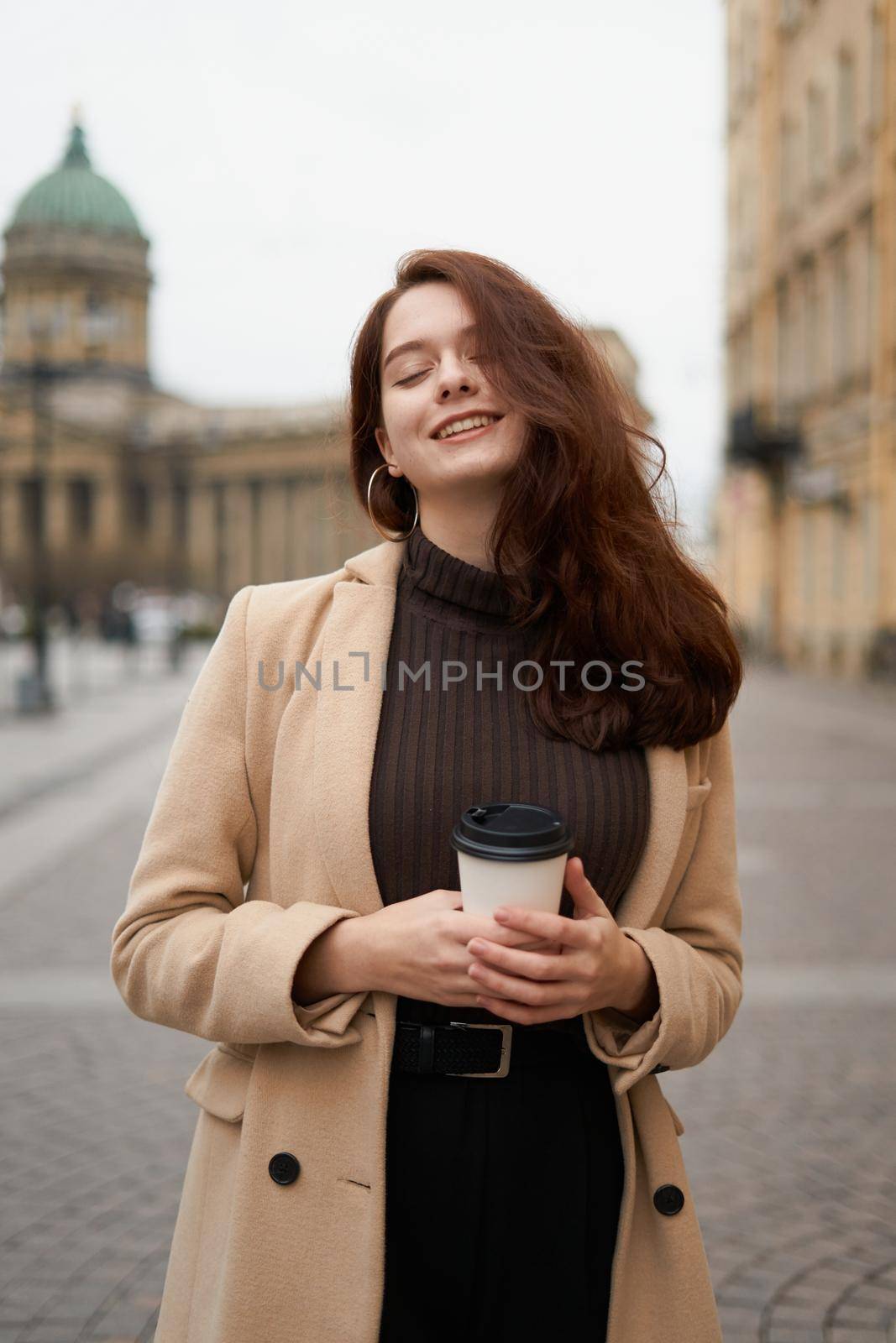 Beautiful serious stylish fashionable smart girl holding cup of coffee in hands and smiles, goes walking down street of St. Petersburg city. Charming thoughtful woman with long dark hair by NataBene