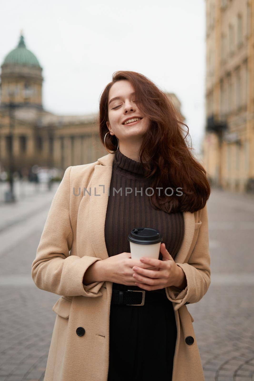 Beautiful serious stylish fashionable smart girl holding cup of coffee in hands and smiles, goes walking down street of St. Petersburg in city center. Charming thoughtful woman with long dark hair