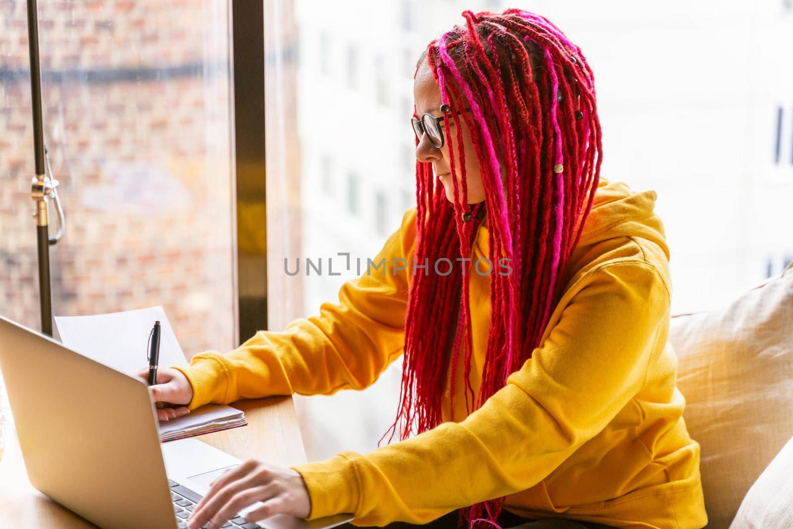 A girl with pink long hair, dreadlocks works at computer in a cafe. The concept of remote work, freelance, digital nomad . Designer is planning design project
