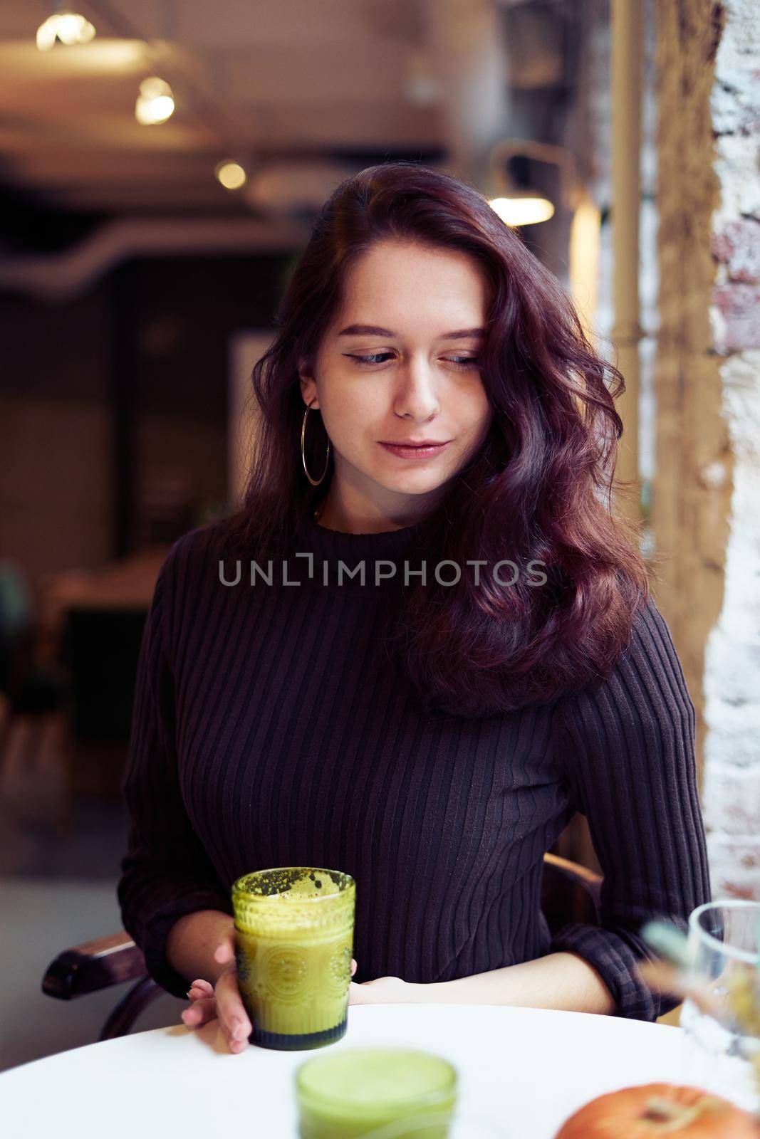 Beautiful serious stylish fashionable smart girl is sitting in cafe and drinking healthy green yellow smoothie or latte vegan. Charming thoughtful woman with long dark brown hair. by NataBene