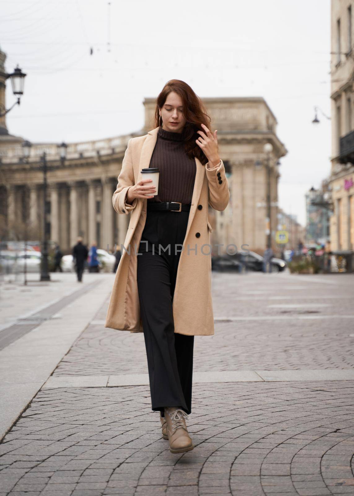 Beautiful serious stylish fashionable smart girl holding cup of coffee in hands goes walking down street of St. Petersburg in city center. Charming thoughtful woman with long dark hair, vertical