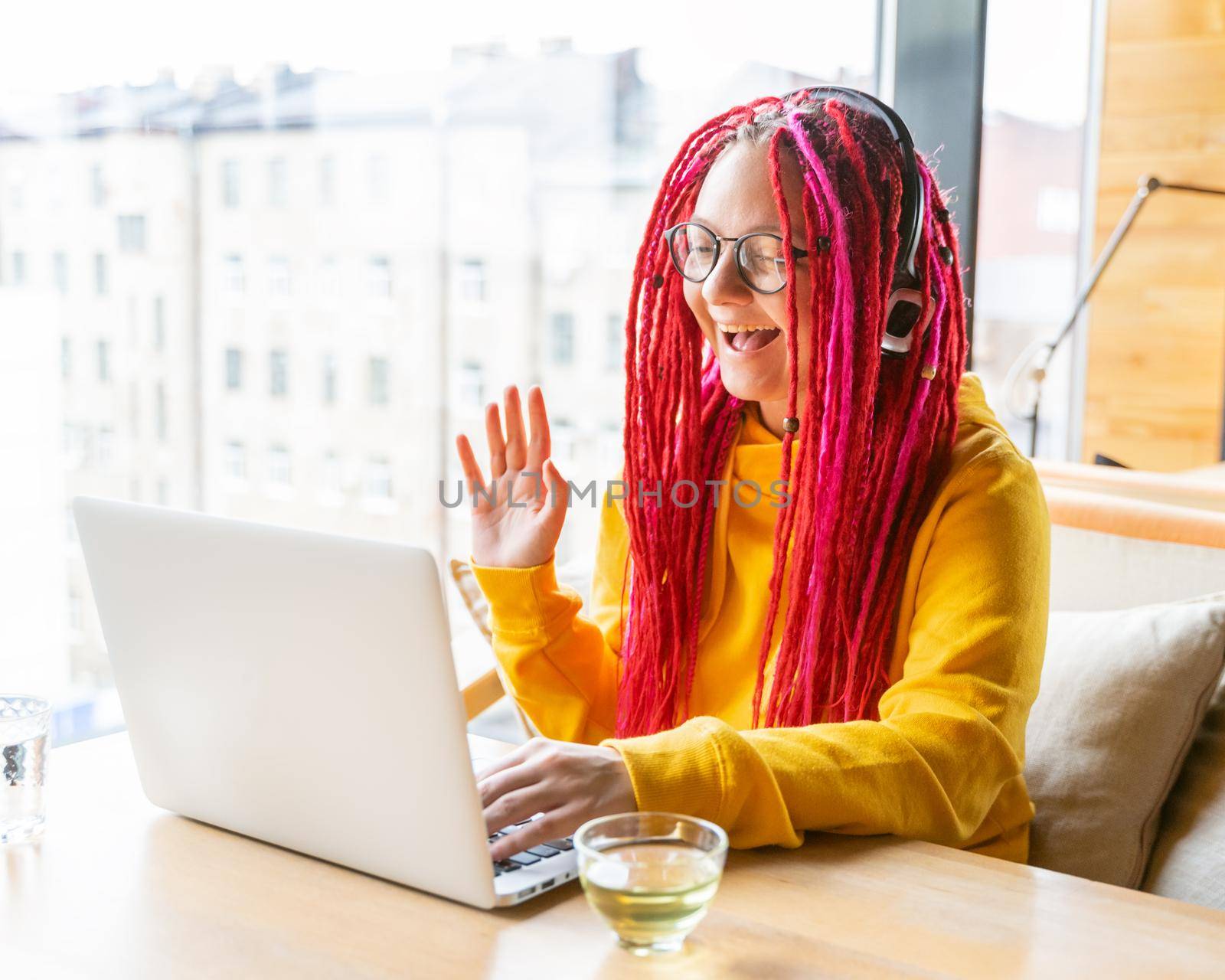 The woman in headphones looks at the laptop, waves her hand and smiles. Live chat, stream. Blogger girl with long pink hair. Good luck concept, positive communication.