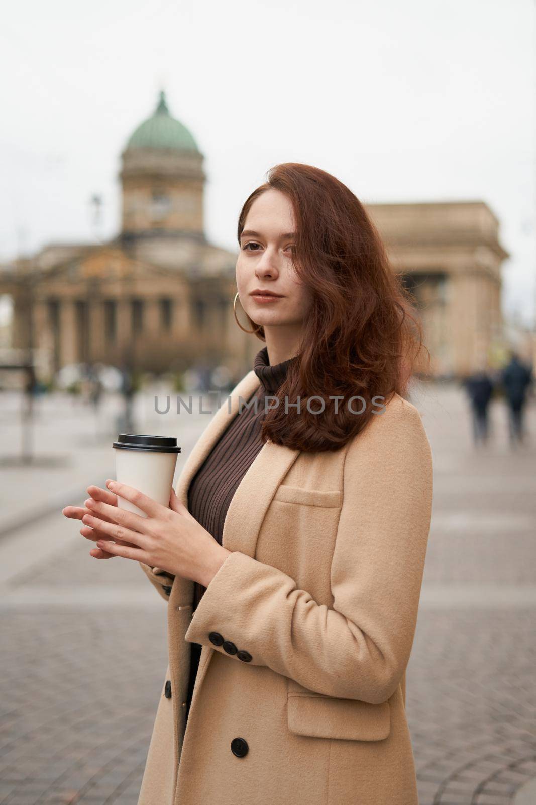 Beautiful serious stylish fashionable smart girl holding cup of coffee in hands goes walking down street of St. Petersburg in city center. Charming thoughtful woman with long dark hair by NataBene