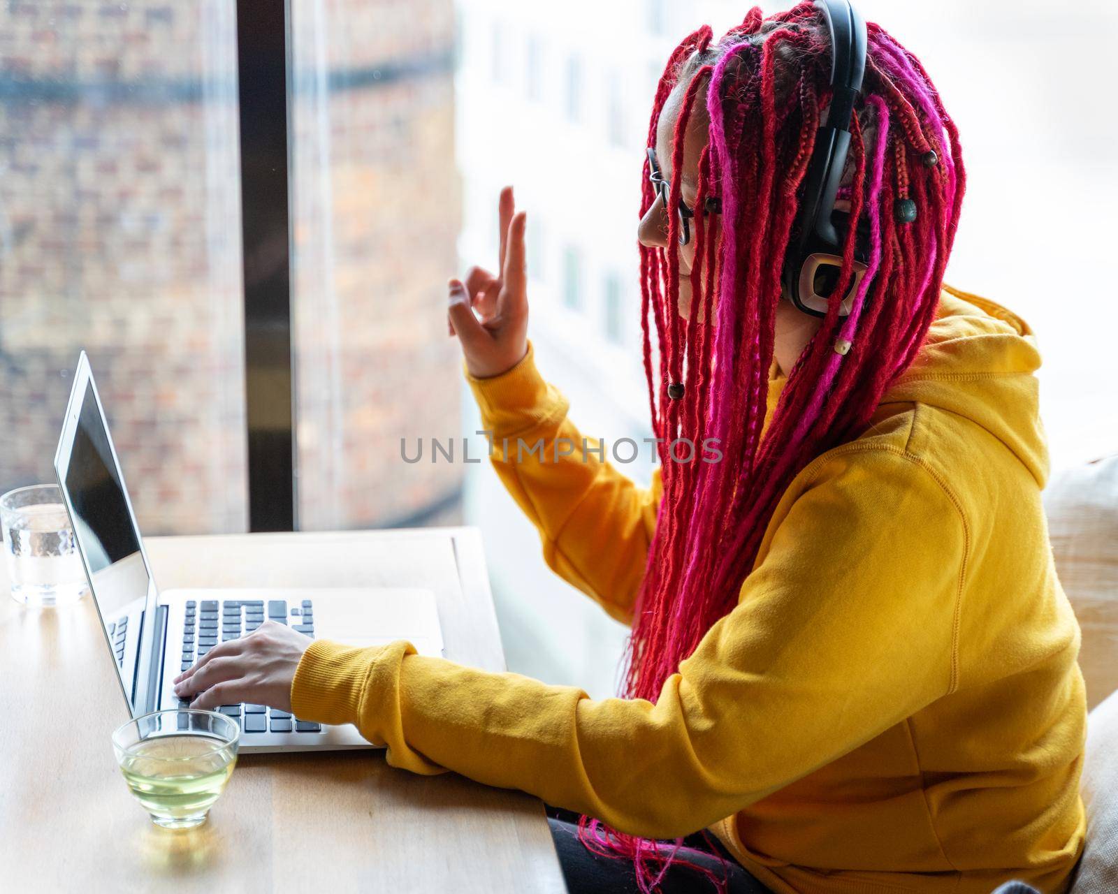 The woman in headphones looks at the laptop and shows a victory sign with her hand. Live chat, stream. Girl with long pink hair, freelancer, blogger. Good luck concept, positive communication.