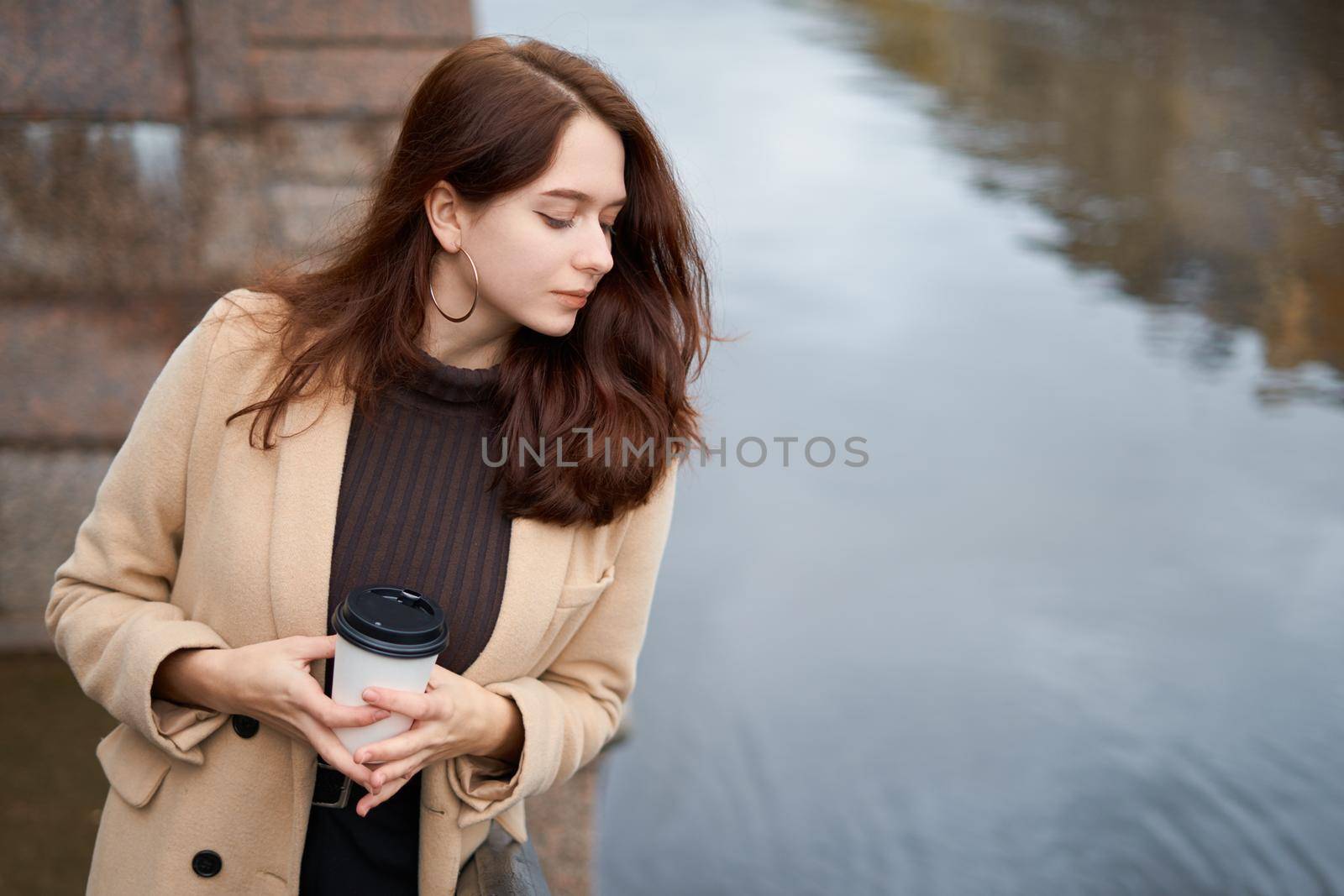 Beautiful serious stylish fashionable girl holding cup of coffee in hands walking street of St. Petersburg in city center. Charming thoughtful woman with long dark hair looks down, copy space