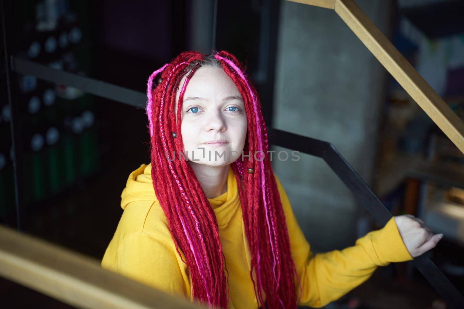 Close-up portrait of a woman with long pink dreadlocks in informal setting, in casual comfortable clothes sitting on the stairs. Beautiful young girl with long hair is sitting in dark cafe