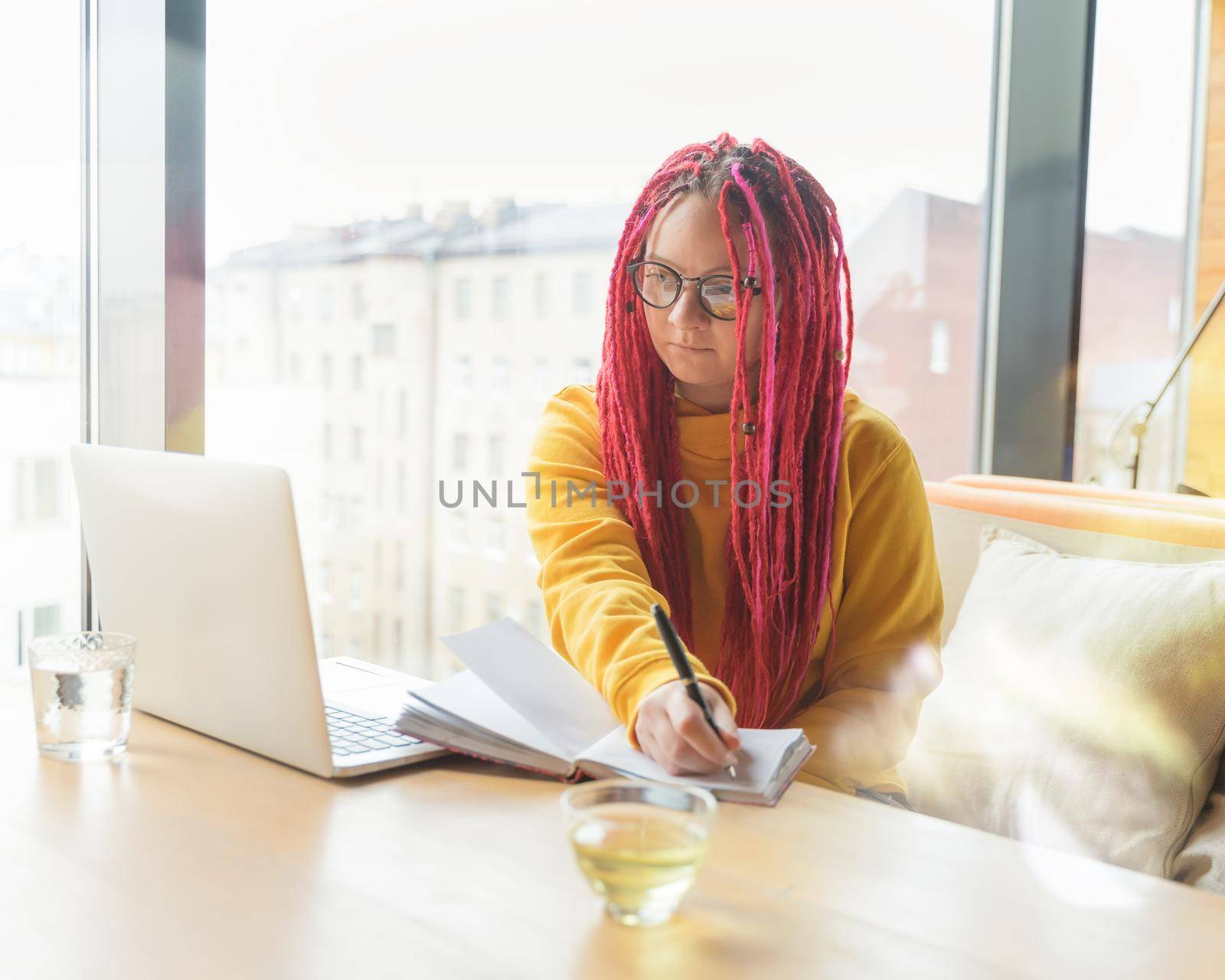 A woman looks into a laptop and writes information in a notebook. Girl with long pink hair, designer, creative specialist, freelancer. Remote Work Concept.