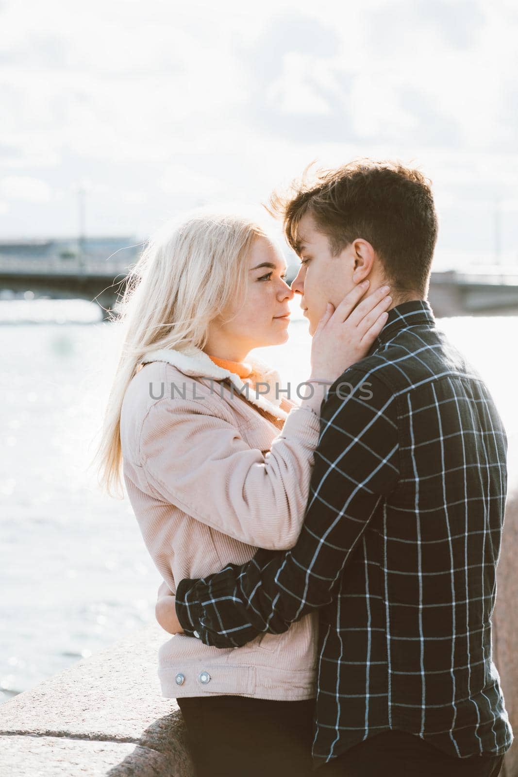 The boy looks tenderly at the girl and wants to kiss. A young couple stands embracing. The concept of teenage love and first kiss, sincere feelings of man and woman. The city, the waterfront. Vertical