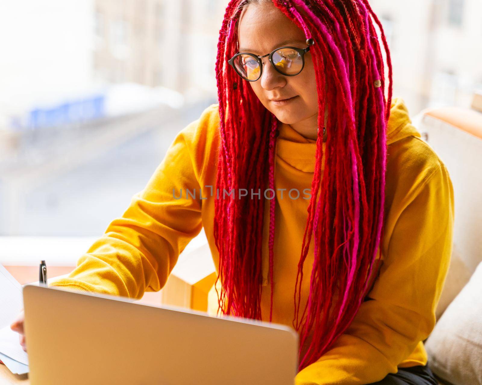 Girl with pink long hair, dreadlocks works at computer in cafe. Concept of remote work, freelance, digital nomad . Designer is planning design project. Desk with laptop, notepad in coworking space