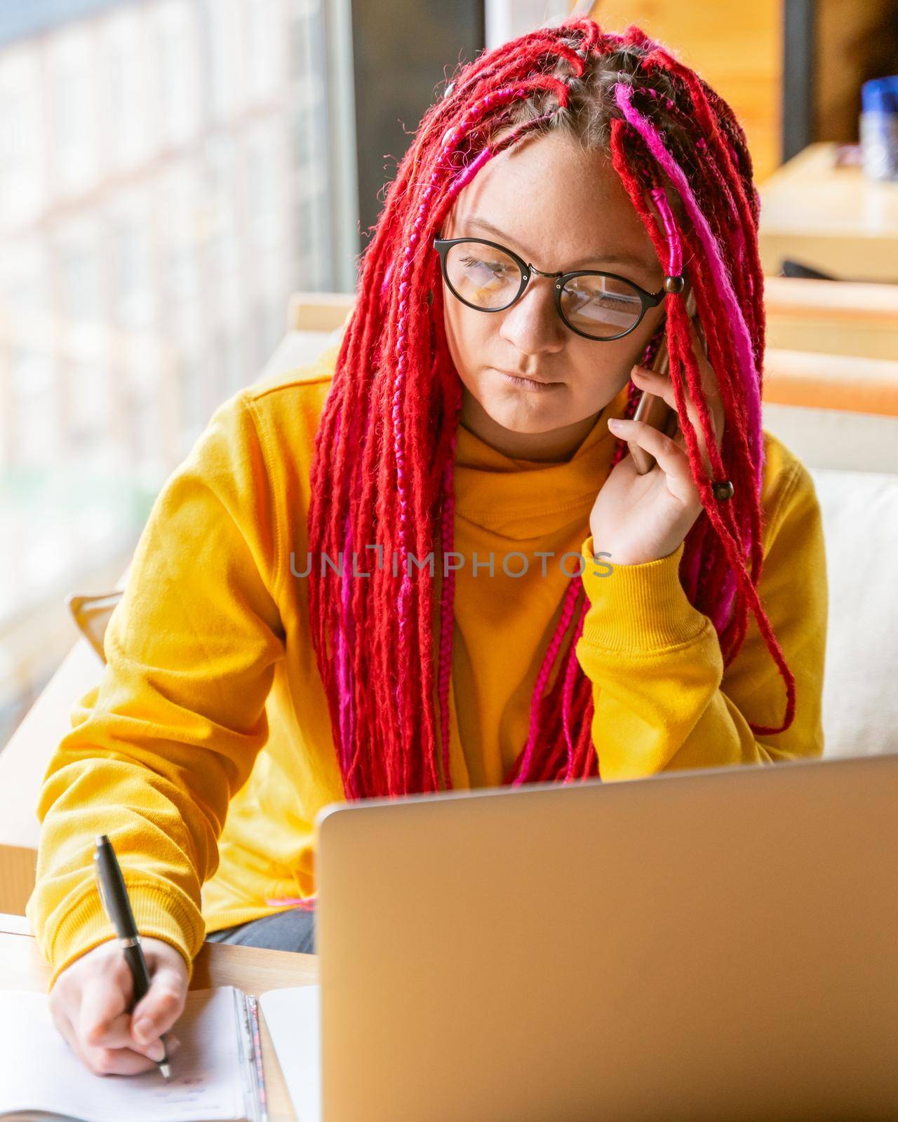 Concept of digital nomad. Girl freelancer talking on mobile phone, working remotely on laptop in cafe, coworking. Woman with long pink dreadlocks in informal setting, in a casual clothes, vertical
